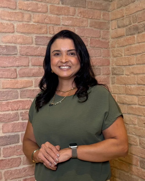 Tricologista Veronica Cardoso sorrindo, de blusa verde, diante de uma parede de tijolinhos